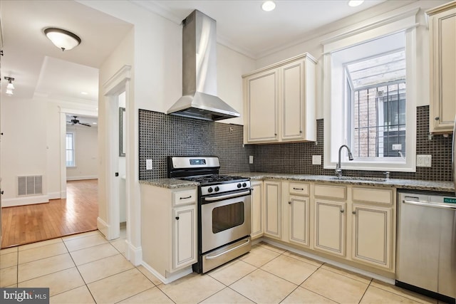 kitchen with wall chimney range hood, stainless steel appliances, sink, light tile patterned floors, and cream cabinetry