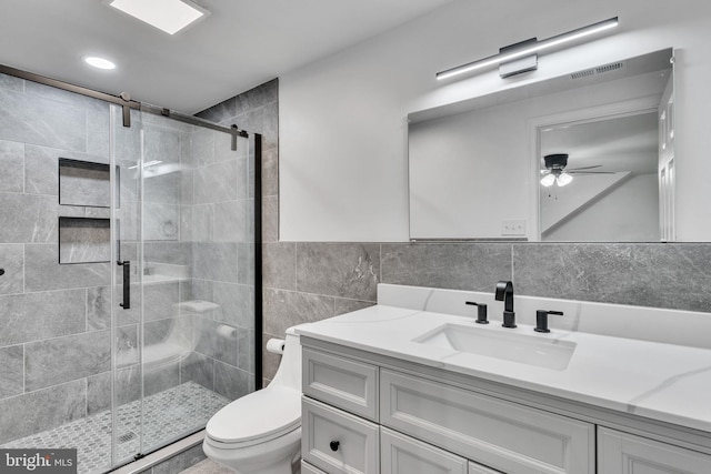 bathroom with toilet, tile walls, a shower with door, ceiling fan, and vanity