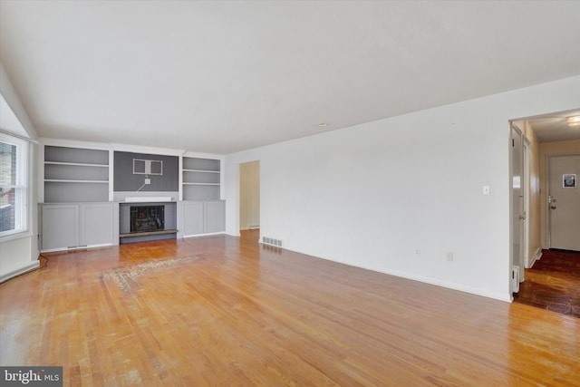 unfurnished living room with built in shelves, a wall mounted air conditioner, a brick fireplace, and light hardwood / wood-style floors