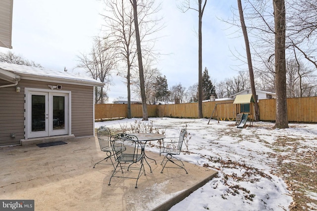 snow covered patio featuring french doors, outdoor dining area, a playground, and a fenced backyard