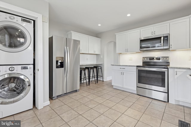 kitchen featuring light stone countertops, stacked washing maching and dryer, light tile patterned floors, white cabinets, and appliances with stainless steel finishes