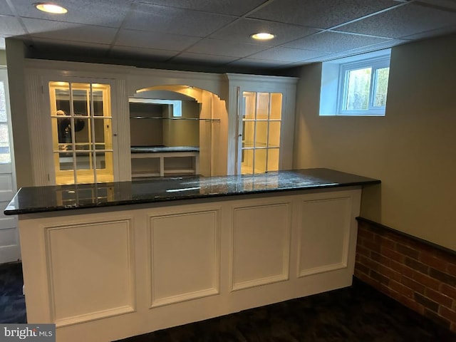 bar with brick wall, white cabinetry, dark stone countertops, and a drop ceiling