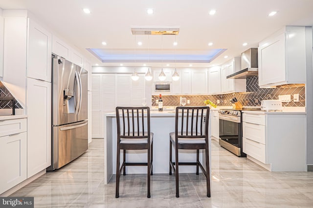 kitchen with hanging light fixtures, a raised ceiling, appliances with stainless steel finishes, white cabinetry, and wall chimney range hood