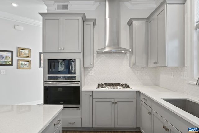 kitchen with stainless steel appliances, gray cabinetry, decorative backsplash, wall chimney range hood, and crown molding