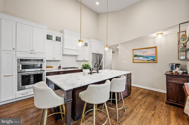 kitchen with appliances with stainless steel finishes, decorative light fixtures, white cabinets, light stone countertops, and a center island with sink