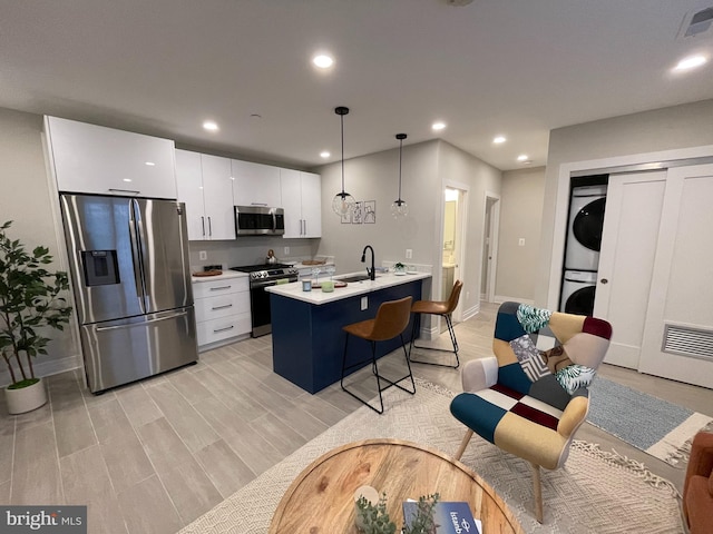 kitchen with white cabinetry, stainless steel appliances, an island with sink, hanging light fixtures, and stacked washing maching and dryer