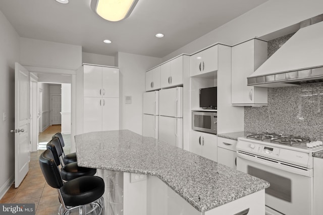 kitchen featuring tasteful backsplash, white appliances, wall chimney exhaust hood, white cabinets, and light stone counters