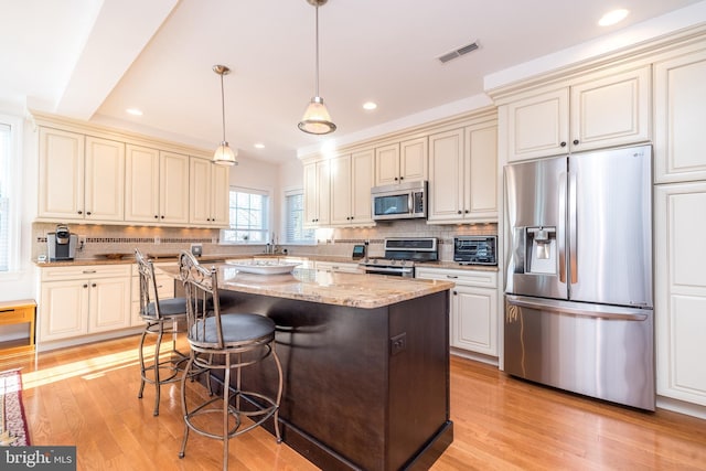 kitchen featuring stainless steel appliances, light stone countertops, pendant lighting, cream cabinets, and a center island