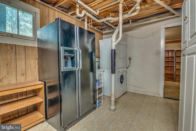 basement featuring stacked washer / drying machine and black refrigerator with ice dispenser