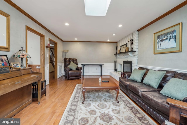 living room featuring a skylight, a wainscoted wall, crown molding, a brick fireplace, and light wood-type flooring