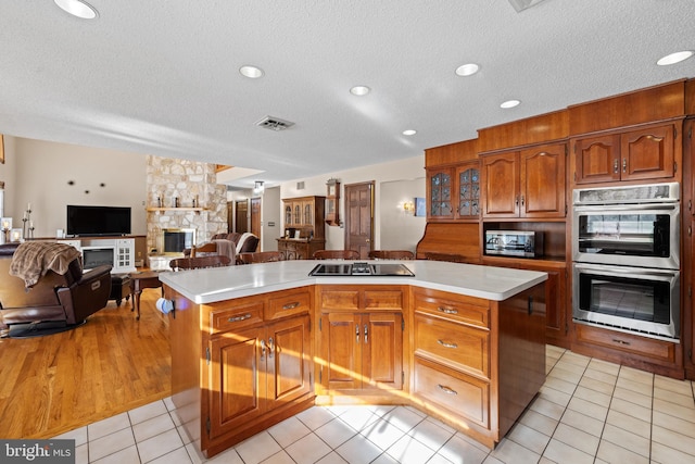 kitchen with stainless steel double oven, a kitchen island, black electric stovetop, a fireplace, and light tile patterned floors