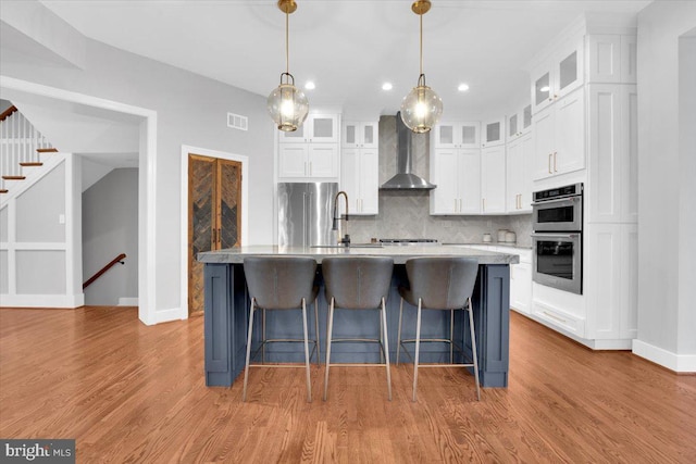 kitchen featuring light wood finished floors, decorative backsplash, stainless steel appliances, white cabinetry, and wall chimney exhaust hood