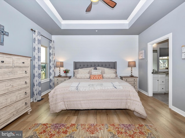 bedroom with light wood-type flooring, ensuite bath, a tray ceiling, and ceiling fan