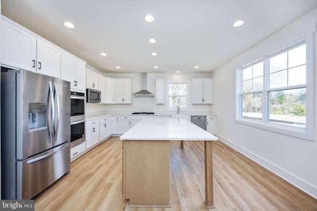 kitchen featuring white cabinetry, a center island, light wood-type flooring, appliances with stainless steel finishes, and wall chimney range hood