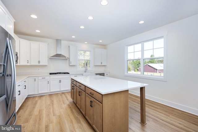 kitchen with wall chimney exhaust hood, light wood-type flooring, appliances with stainless steel finishes, a kitchen island, and white cabinets