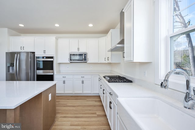 kitchen with wall chimney exhaust hood, sink, white cabinetry, light wood-type flooring, and stainless steel appliances