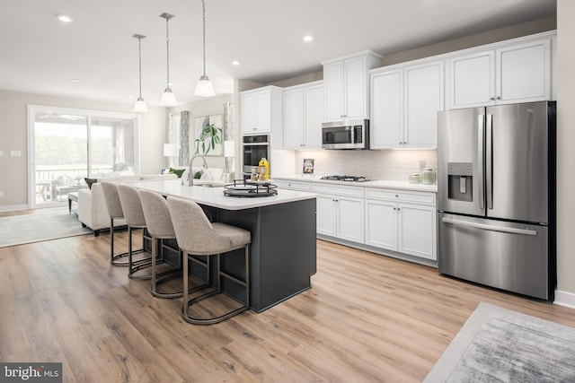 kitchen featuring white cabinetry, stainless steel appliances, decorative light fixtures, light wood-type flooring, and a kitchen island with sink