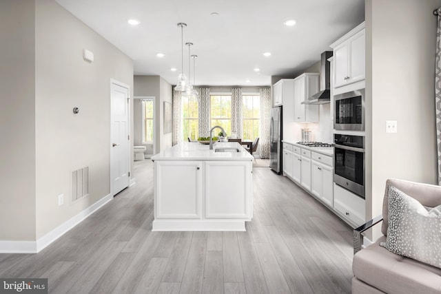 kitchen featuring wall chimney range hood, pendant lighting, a kitchen island with sink, stainless steel appliances, and white cabinets