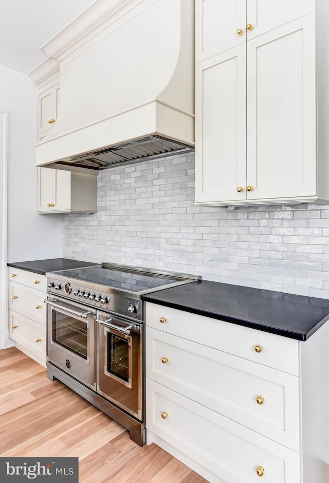 kitchen with white cabinetry, custom exhaust hood, backsplash, range with two ovens, and light hardwood / wood-style flooring