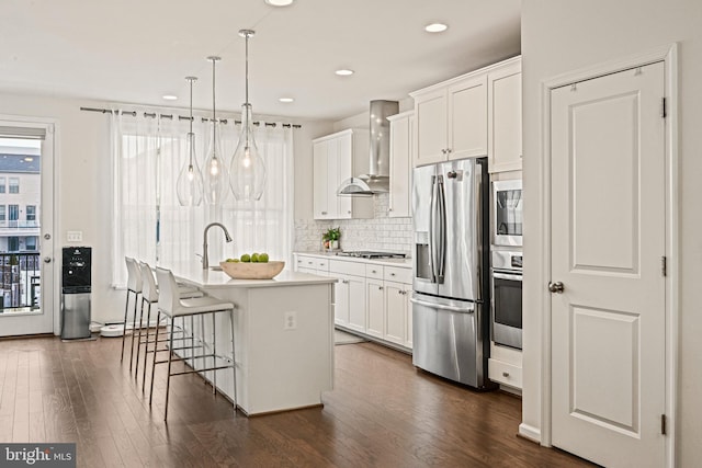 kitchen with wall chimney exhaust hood, white cabinetry, an island with sink, pendant lighting, and stainless steel appliances