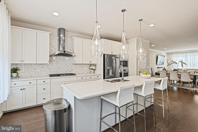 kitchen with wall chimney range hood, stainless steel appliances, white cabinets, a center island with sink, and decorative light fixtures