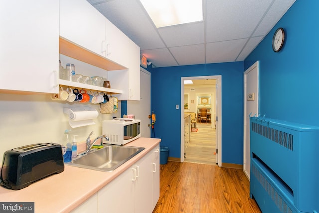 kitchen featuring light wood-type flooring, a paneled ceiling, heating unit, white cabinets, and sink