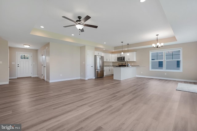 unfurnished living room with a raised ceiling, light wood-type flooring, sink, and ceiling fan with notable chandelier