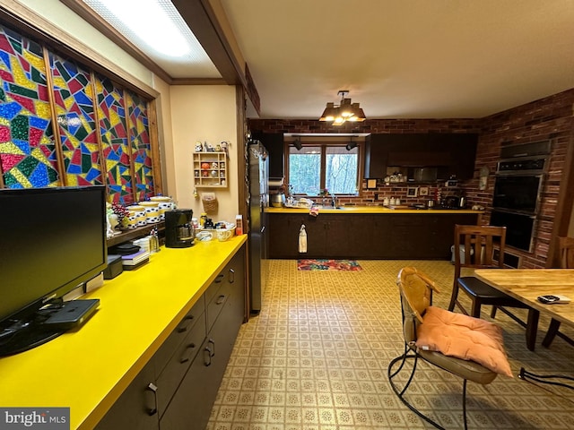 kitchen featuring dark brown cabinetry, an inviting chandelier, crown molding, double wall oven, and brick wall