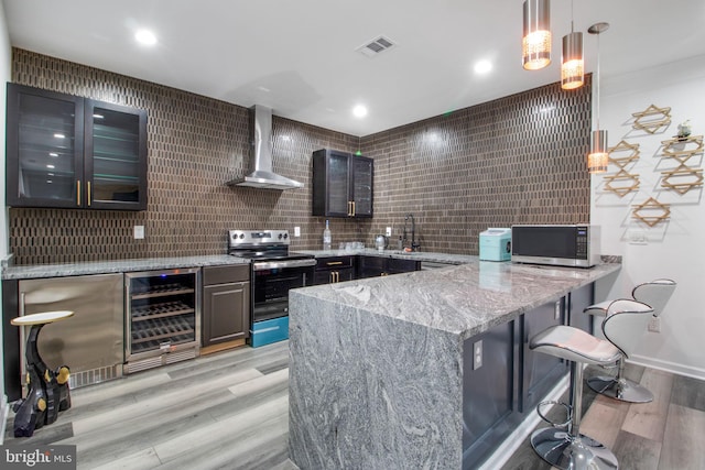 kitchen with wine cooler, light hardwood / wood-style flooring, hanging light fixtures, appliances with stainless steel finishes, and wall chimney range hood