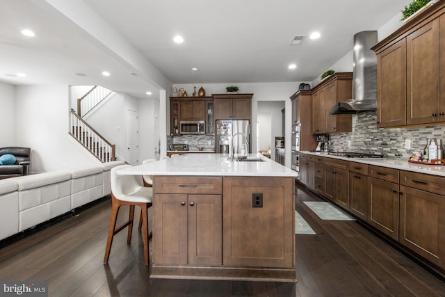 kitchen featuring wall chimney range hood, sink, dark wood-type flooring, appliances with stainless steel finishes, and a kitchen island with sink