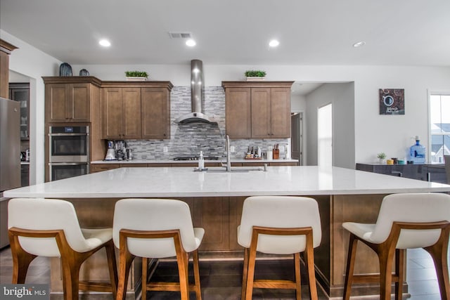 kitchen with sink, decorative backsplash, a large island with sink, stainless steel double oven, and wall chimney exhaust hood