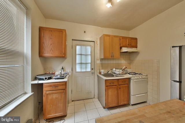 kitchen featuring backsplash, white gas range oven, sink, stainless steel refrigerator, and light tile patterned floors