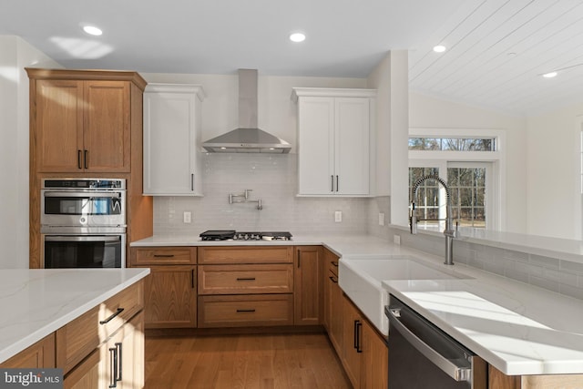 kitchen featuring light stone counters, stainless steel appliances, light wood-style flooring, a sink, and wall chimney range hood