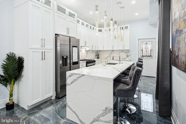 kitchen featuring light stone countertops, decorative light fixtures, white cabinetry, an island with sink, and stainless steel fridge