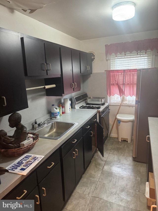 kitchen featuring freestanding refrigerator, dark cabinetry, a sink, and gas range oven