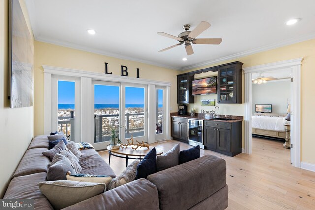 living room with beverage cooler, ceiling fan, crown molding, wet bar, and light wood-type flooring