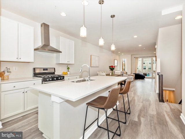 kitchen featuring white cabinets, wall chimney range hood, sink, a center island with sink, and stainless steel range with gas stovetop