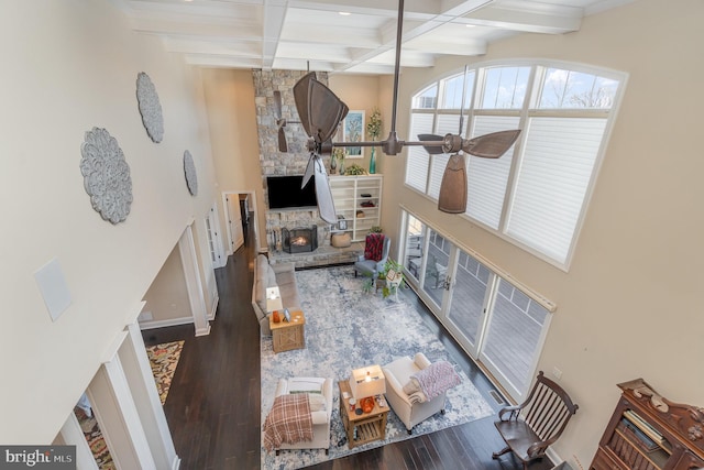 living room with ceiling fan, beamed ceiling, coffered ceiling, dark wood-type flooring, and a stone fireplace