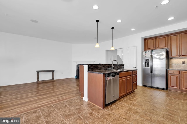 kitchen with dark stone countertops, hanging light fixtures, a kitchen island with sink, and stainless steel appliances