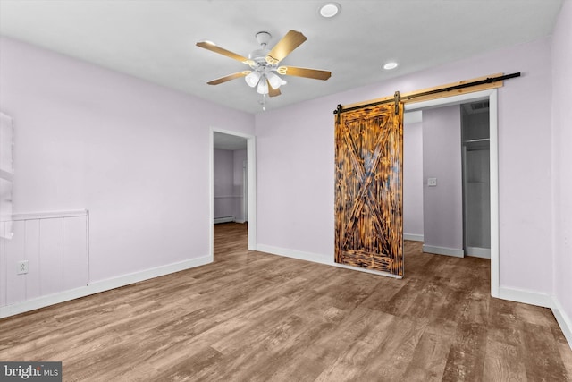 unfurnished bedroom with baseboard heating, a barn door, hardwood / wood-style floors, and ceiling fan