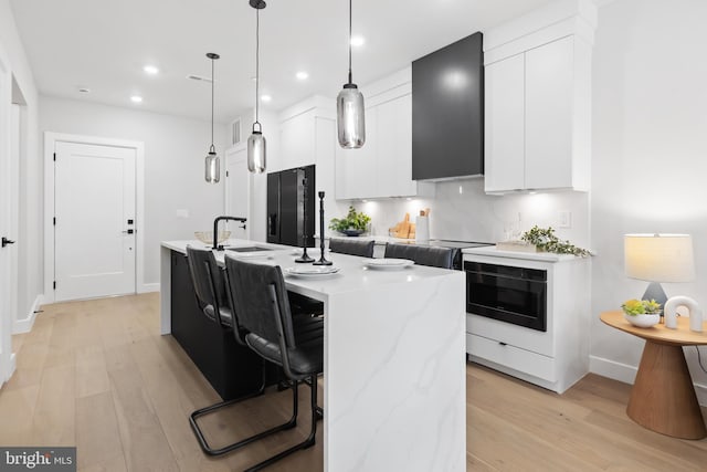 kitchen featuring white cabinets, decorative light fixtures, an island with sink, light stone counters, and range hood