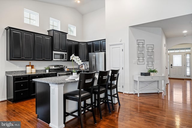 kitchen featuring dark hardwood / wood-style flooring, appliances with stainless steel finishes, light stone countertops, a kitchen bar, and a kitchen island with sink