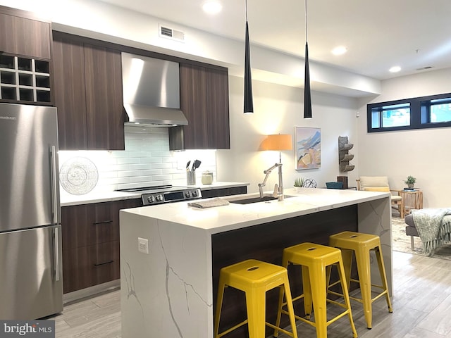 kitchen featuring wall chimney range hood, stainless steel fridge, a kitchen island with sink, a kitchen bar, and decorative backsplash