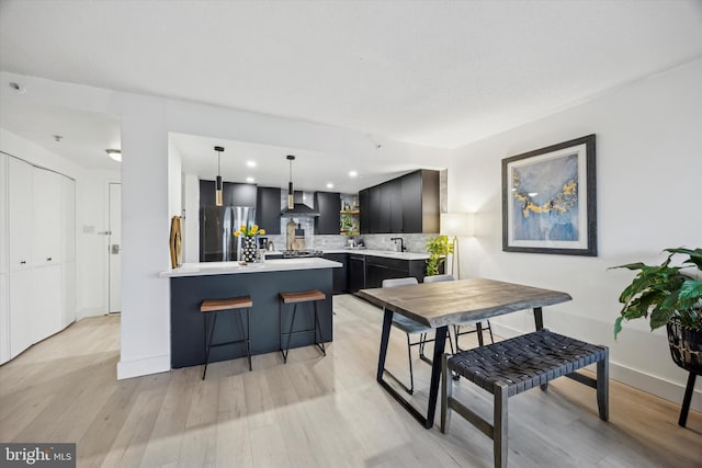 kitchen featuring decorative light fixtures, tasteful backsplash, wall chimney range hood, light hardwood / wood-style floors, and stainless steel refrigerator