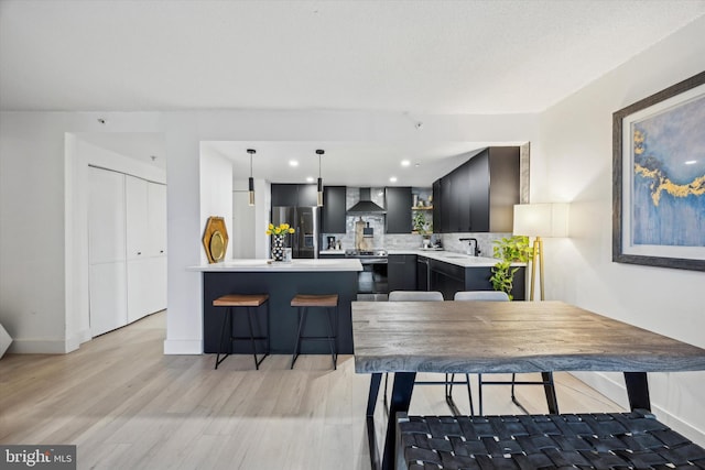 kitchen featuring appliances with stainless steel finishes, wall chimney exhaust hood, light hardwood / wood-style floors, hanging light fixtures, and a breakfast bar area