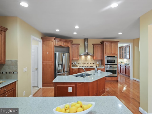 kitchen with appliances with stainless steel finishes, an island with sink, sink, wall chimney range hood, and light hardwood / wood-style flooring