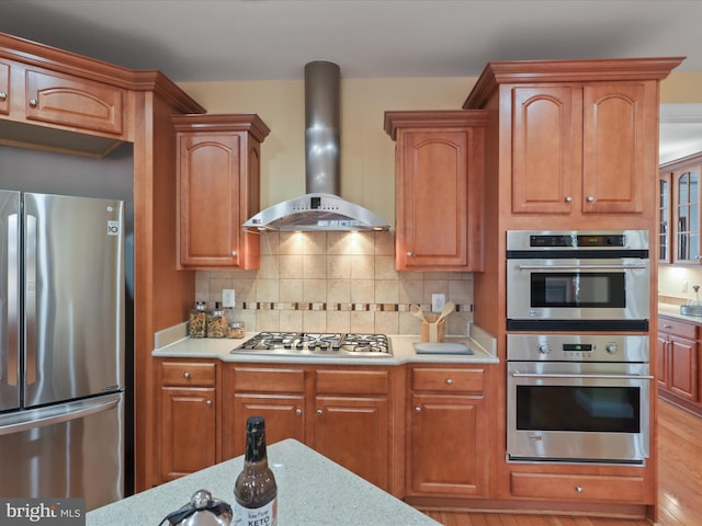 kitchen with appliances with stainless steel finishes, backsplash, light stone counters, wall chimney range hood, and light wood-type flooring