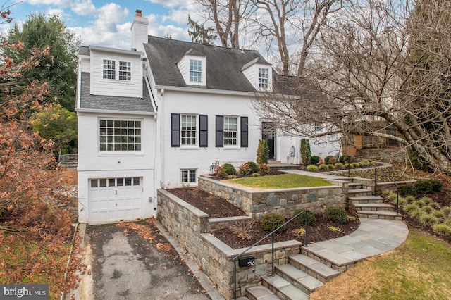 view of front of house featuring aphalt driveway, a garage, brick siding, a shingled roof, and a chimney