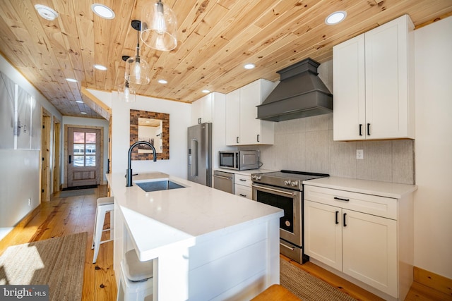 kitchen with an island with sink, stainless steel appliances, custom range hood, pendant lighting, and sink