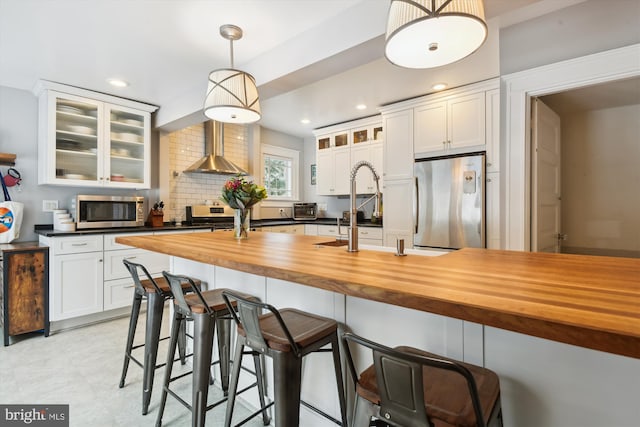 kitchen featuring pendant lighting, white cabinetry, wall chimney range hood, stainless steel appliances, and butcher block countertops